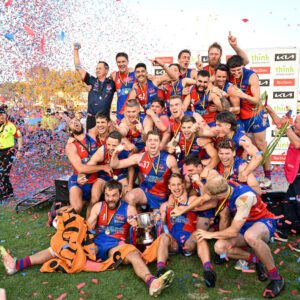 A winning men's football team cheering in a group photo, confetti explodes to their right, while two team players on the ground both grasp the winning cup